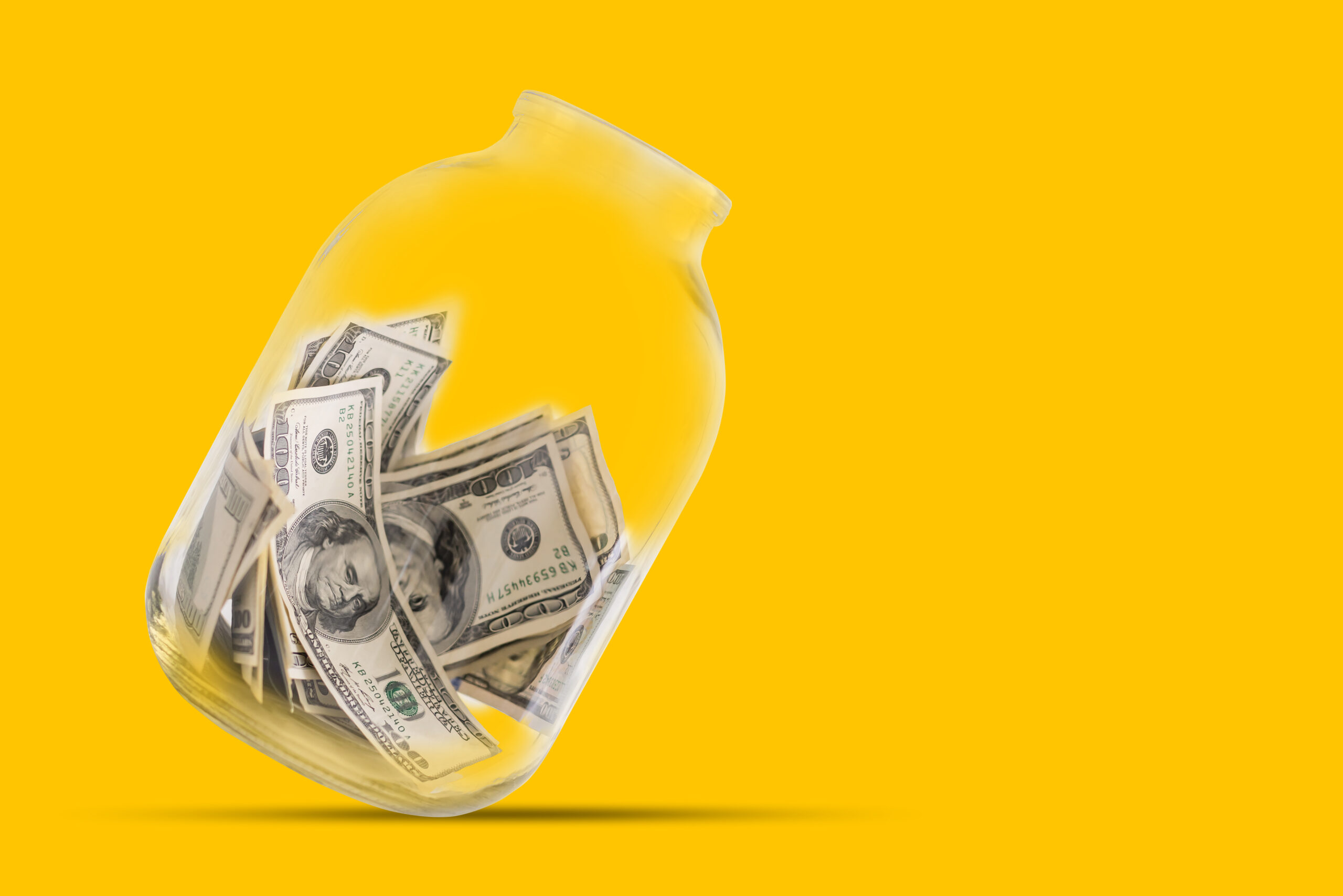 Many 100 US dollars bank notes in a glass jar isolated on yellow background.